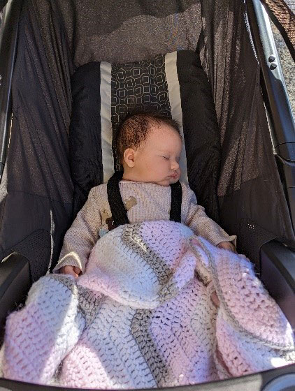 A baby tucked in with a pink, yellow, and gray blanket sleeps peacefully in a stroller.