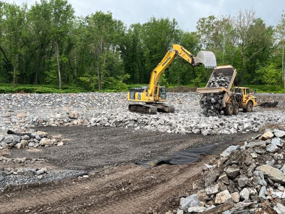 A backhoe and dumptruck layer small and large rocks in a channel surrounded by trees.