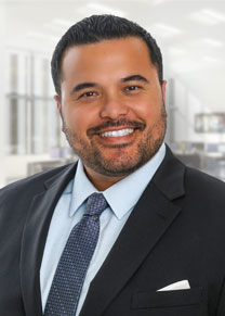 A man wearing a black suit and gray patterned tie smiles for a professional headshot.