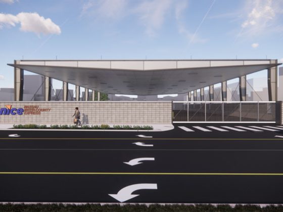 from the street looking at a white battery electric bus charging station building with a white brick wall and pedestrians in the street.