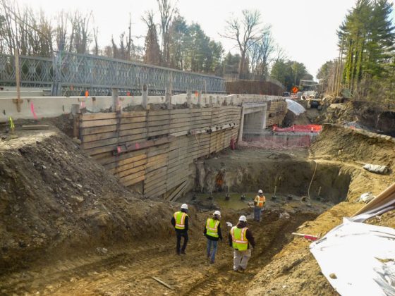 A construction site with workers descending into a pit with wooden shoring below a roadway bridge.