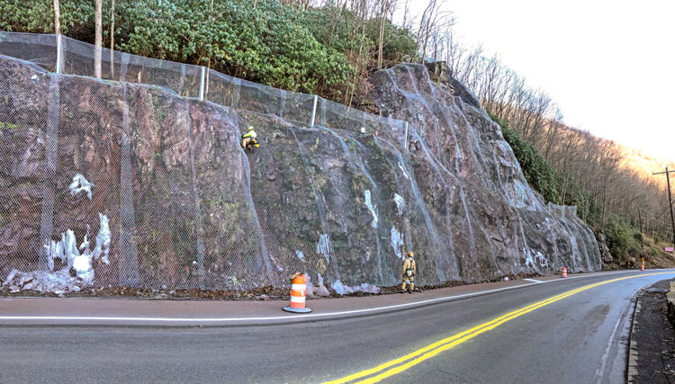 Workers in safety gear work on anchored wire mesh covering a potential rockfall stretch along the edge of a highway.