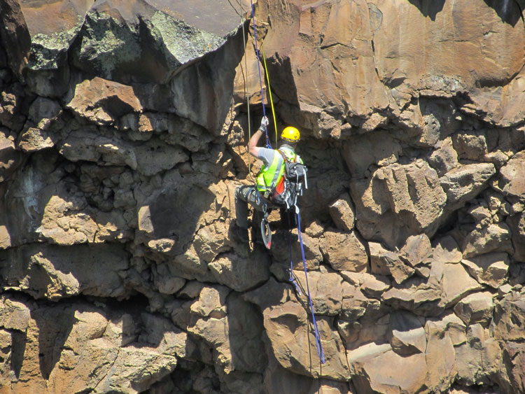 A man in a climbing harness, safety jacket, and a hard hat inspects the side of a rock slope.
