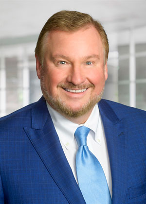 Formal headshot of a white man with light brown hair and beard in a dark blue suit and light blue tie.